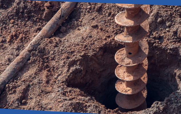 Rusty earth auger drilling into dry soil, with a deep hole and metal rod visible; close-up construction scene conveys rugged, gritty work.